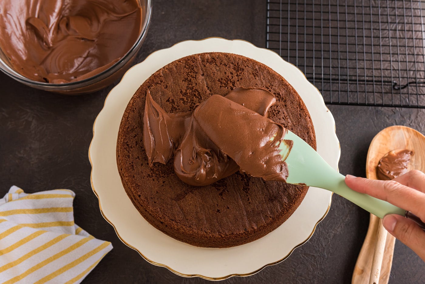spreading chocolate fudge frosting over chocolate cake with a rubber spatula