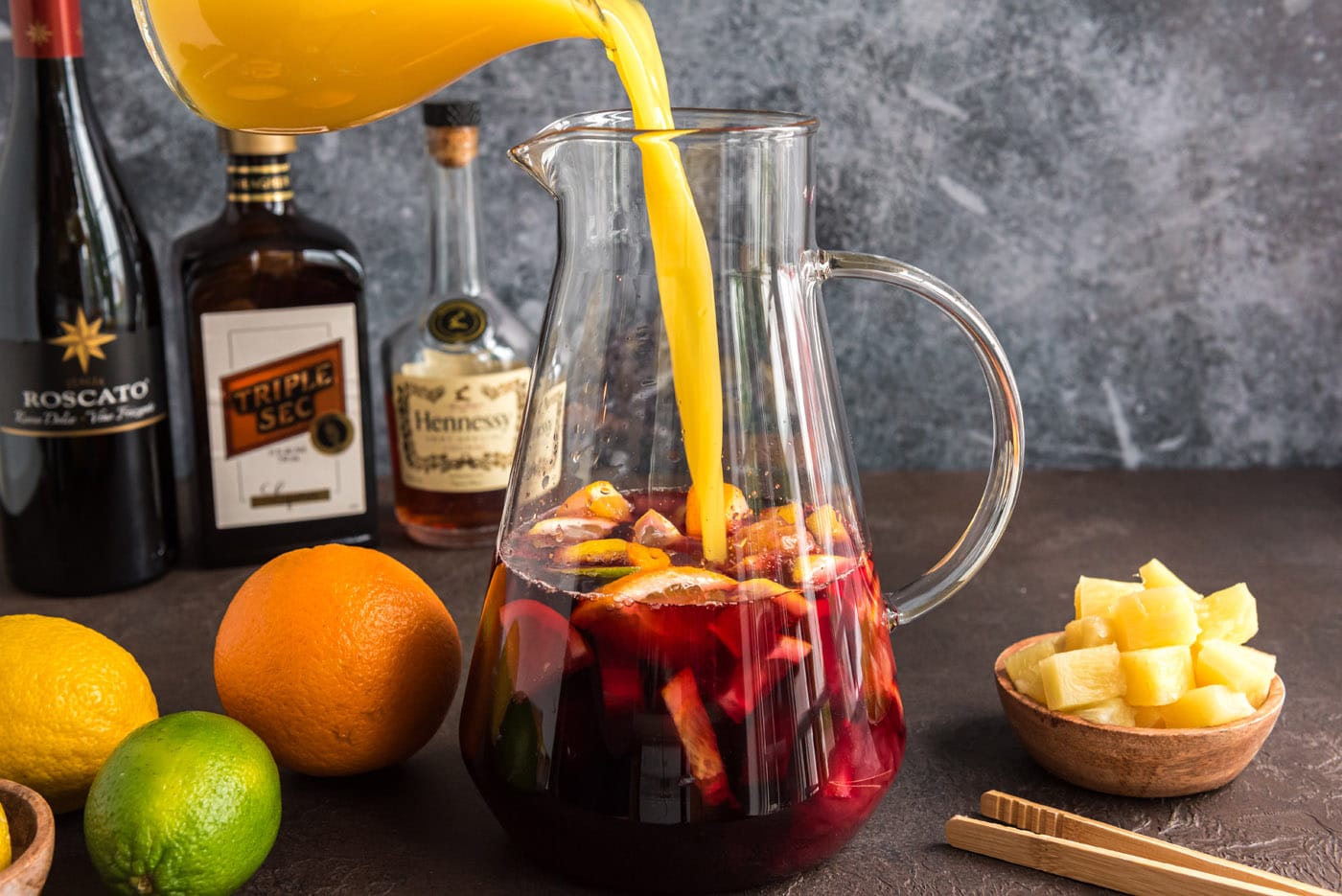 pouring orange juice into pitcher of red wine, brandy, and triple sec