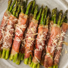 Close up photo of Prosciutto Wrapped Asparagus on a plate topped with shaved parmesan