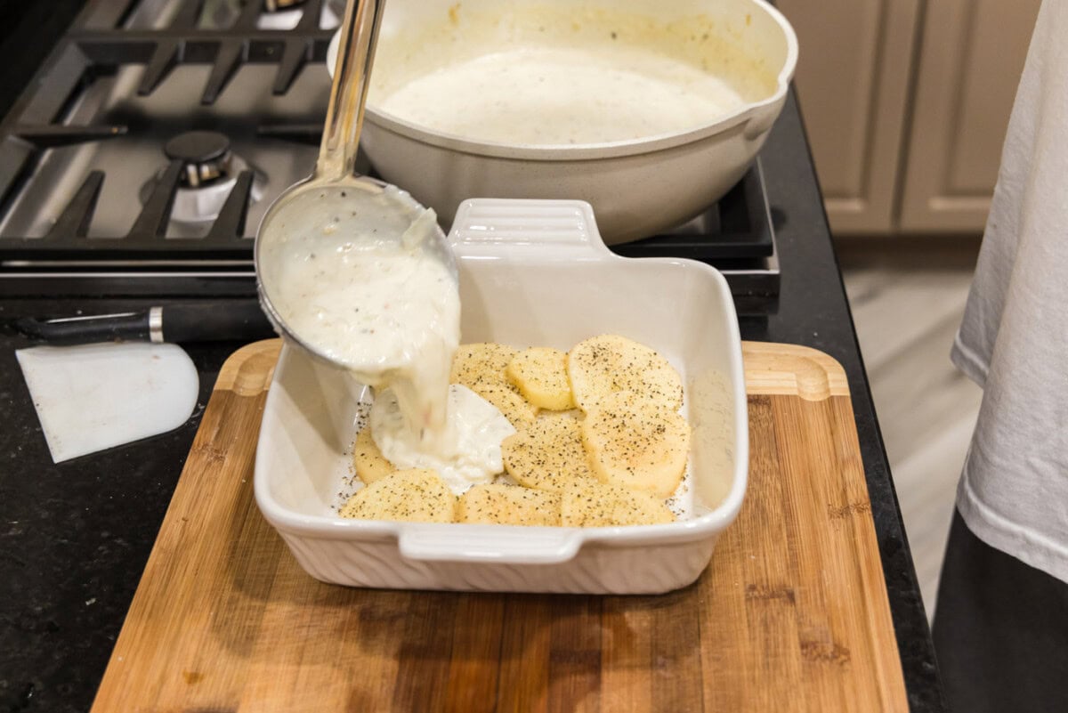 ladeling cream sauce over scalloped potatoes in a dish