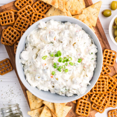 Bowl of Dirty Martini Dip on a board with pretzels and crackers around it