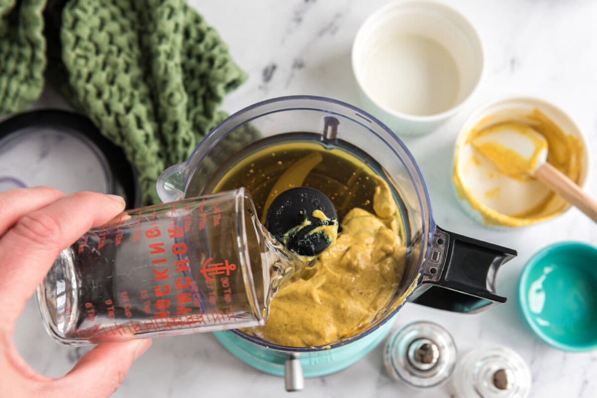 pouring water into food processor with mustard, olive oil, and honey