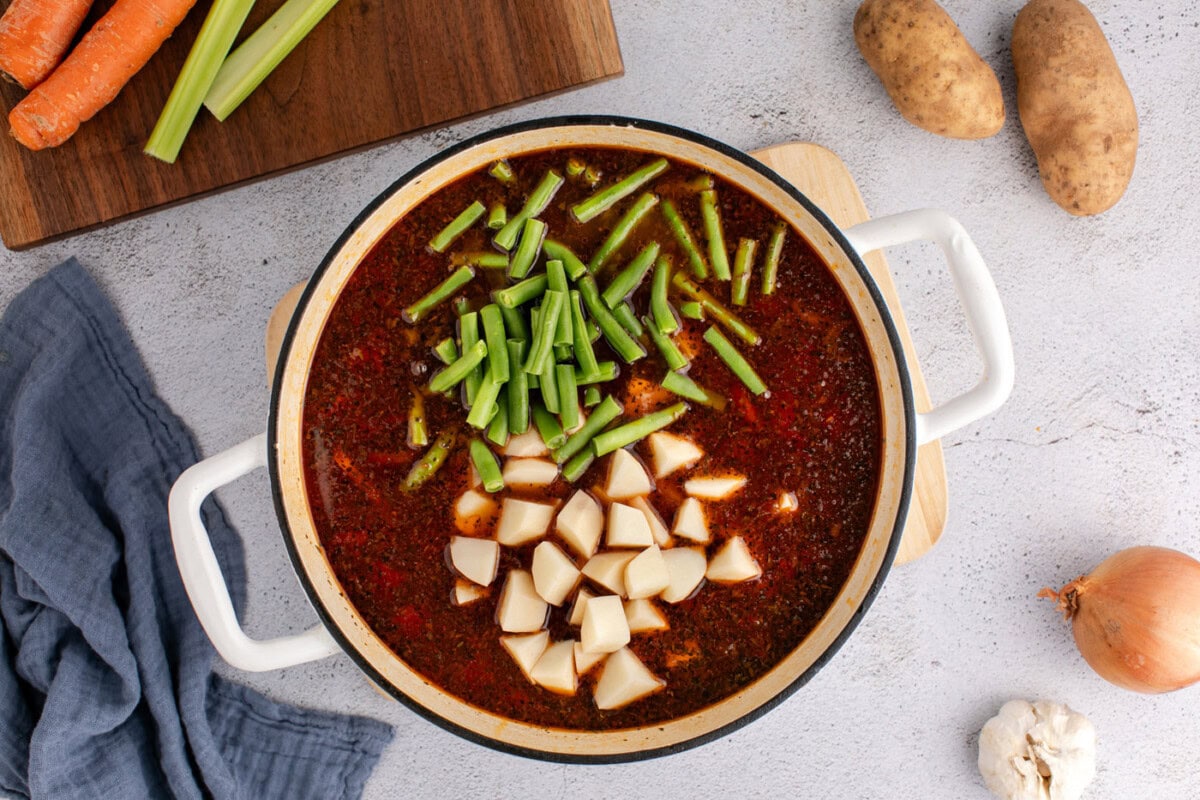 cubed potatoes and trimmed green beans added to vegetable beef soup in pot