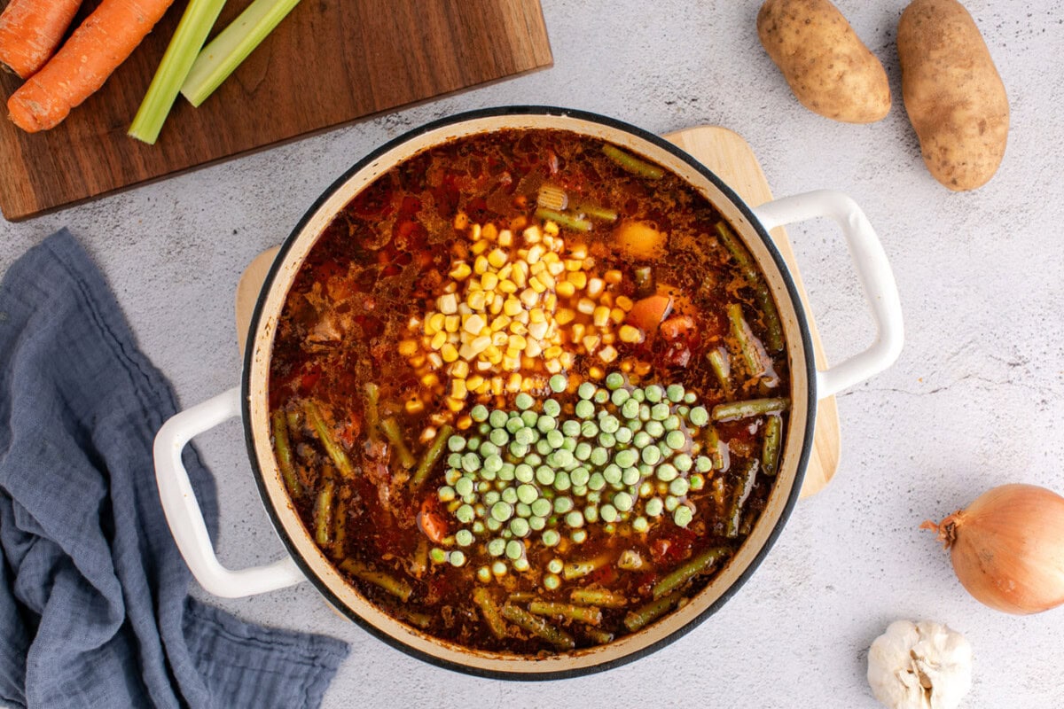 corn and frozen peas added to vegetable beef soup in pot