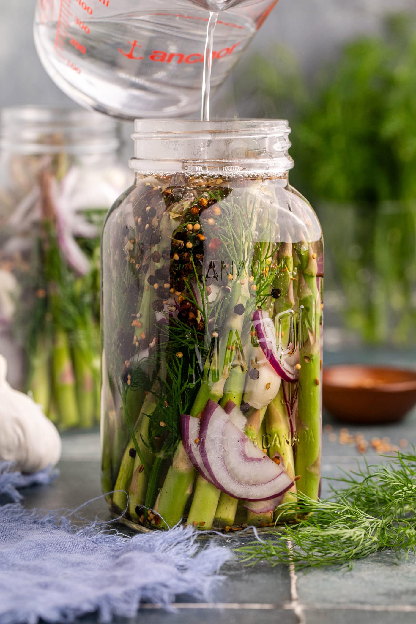 pouring vinegar brine over asparagus, red onion, garlic, and dill