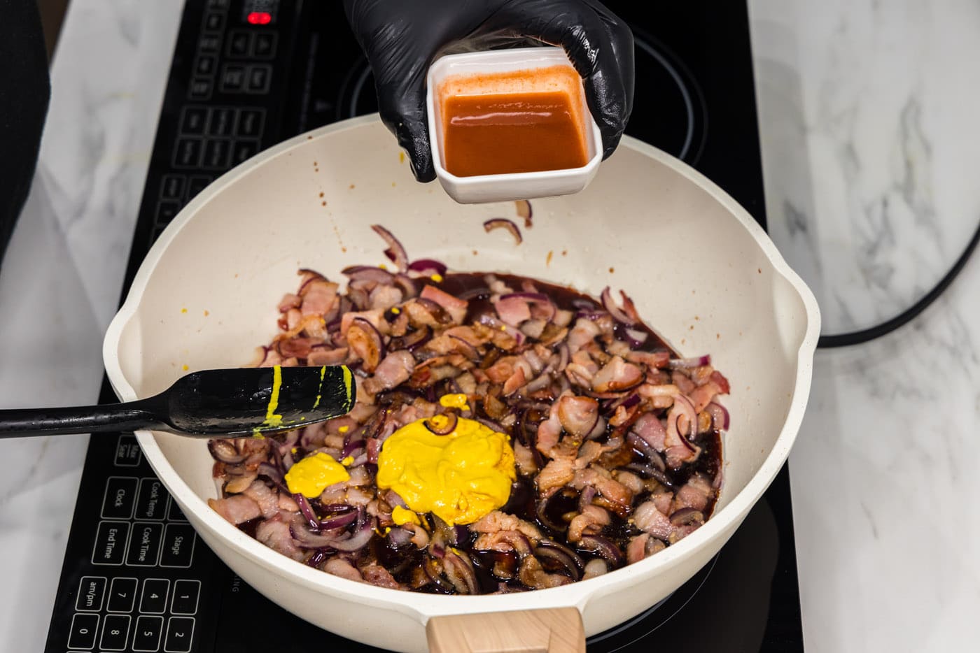 adding hot sauce to mixture of bacon, mustard, onions, and soy sauce in a skillet