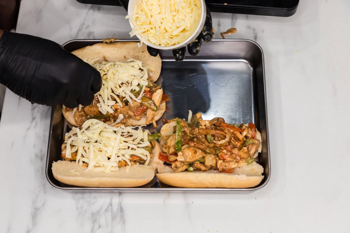sprinkling mozzarella cheese over chicken philly cheesesteak sandwiches