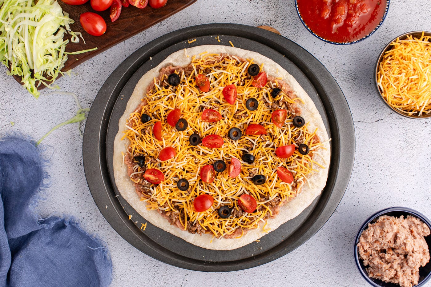 shredded cheese, taco meat, tomatoes, and olives added on top of refried beans and pizza crust