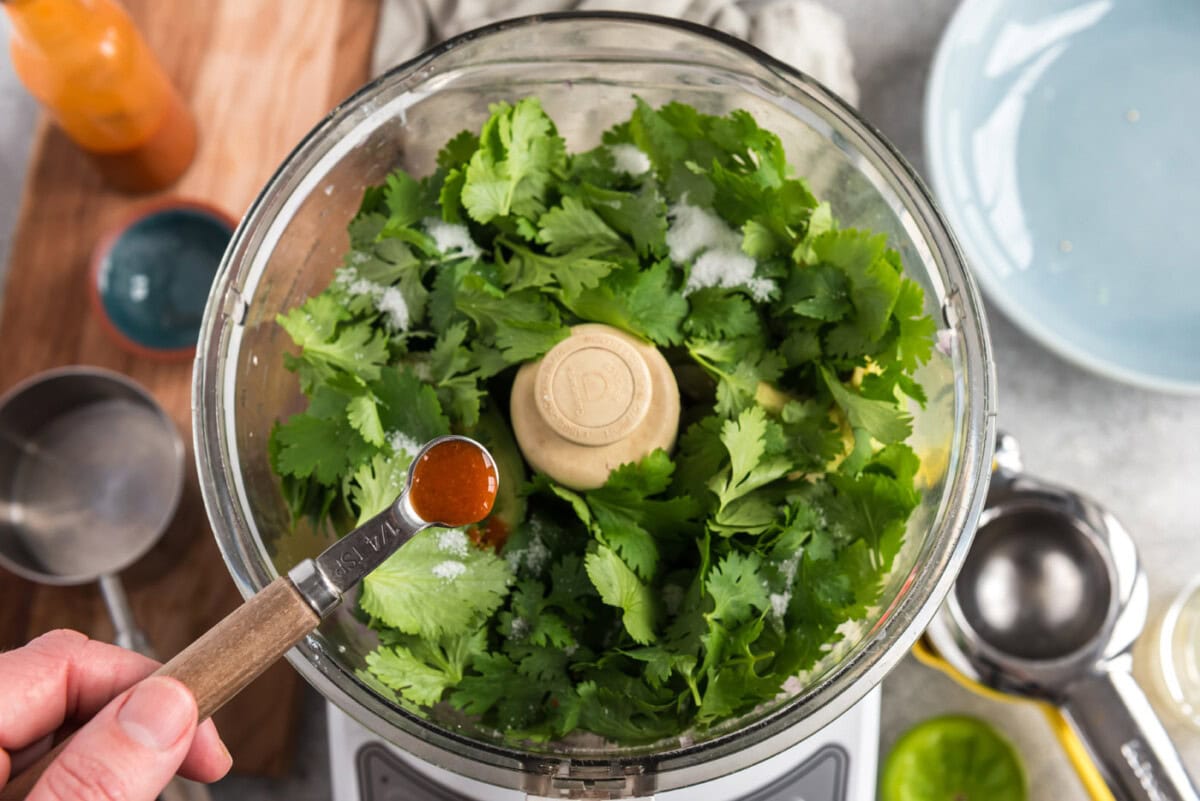 adding hot pepper sauce to food processor with cilantro, onion, avocado, and chile peppers