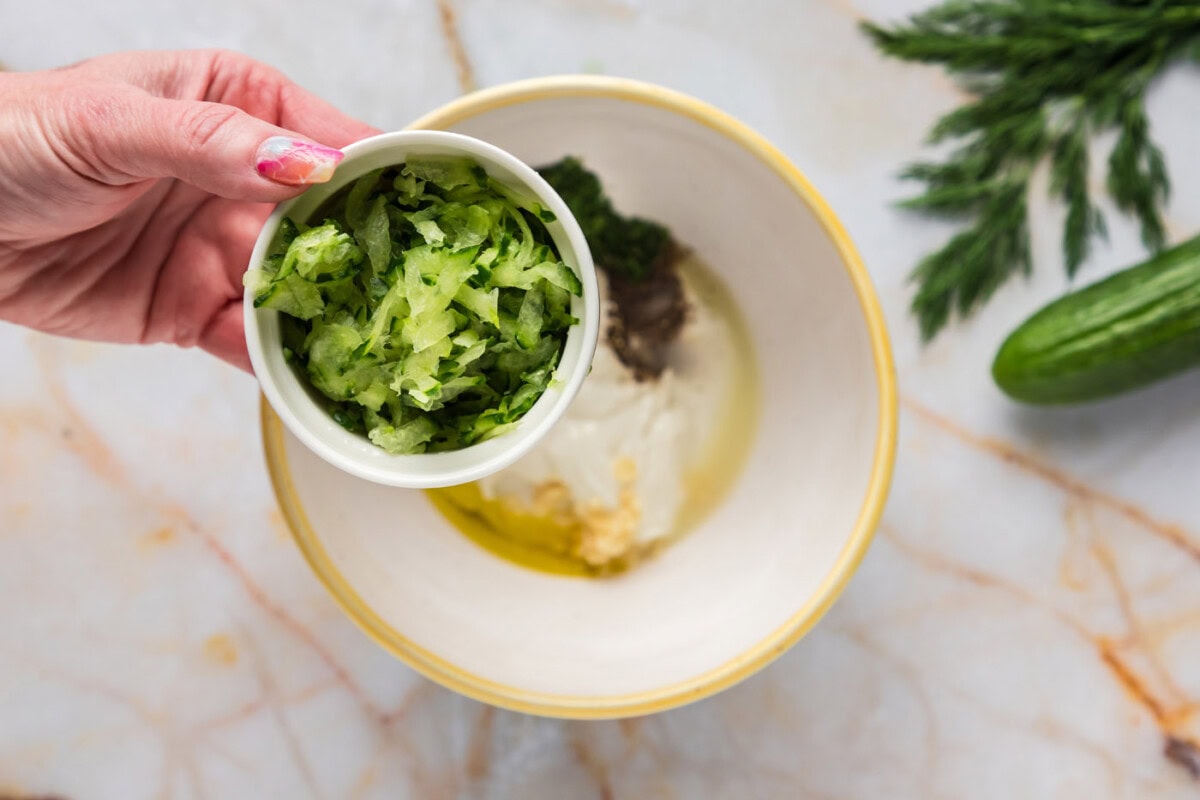 adding shredded cucumber to a bowl of tzatziki sauce ingredients