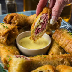 Half a Corned Beef Egg Roll held above a bowl of dipping sauce