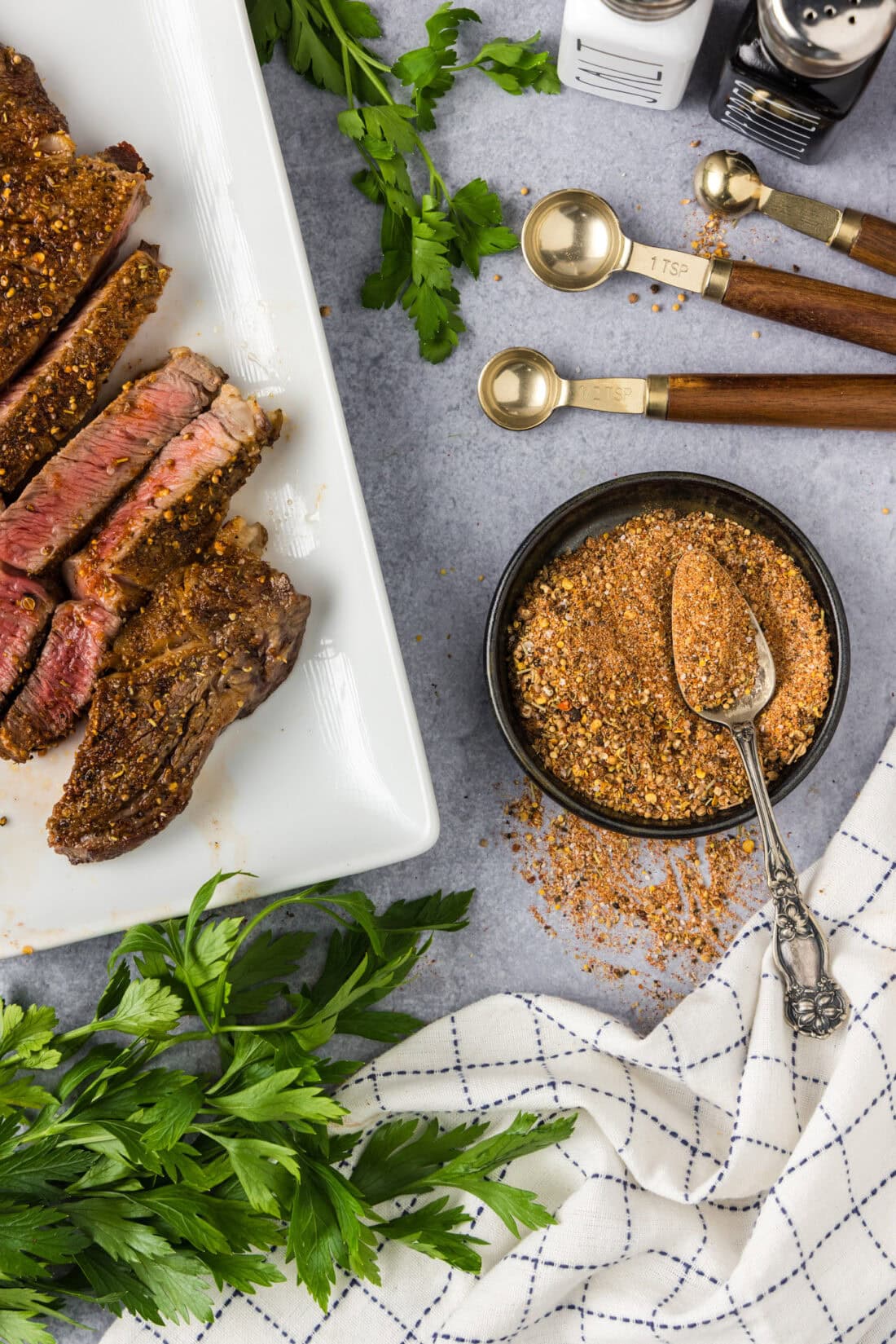 Bowl of Homemade Montreal Steak Seasoning with a spoon in it and cooked steak to the side