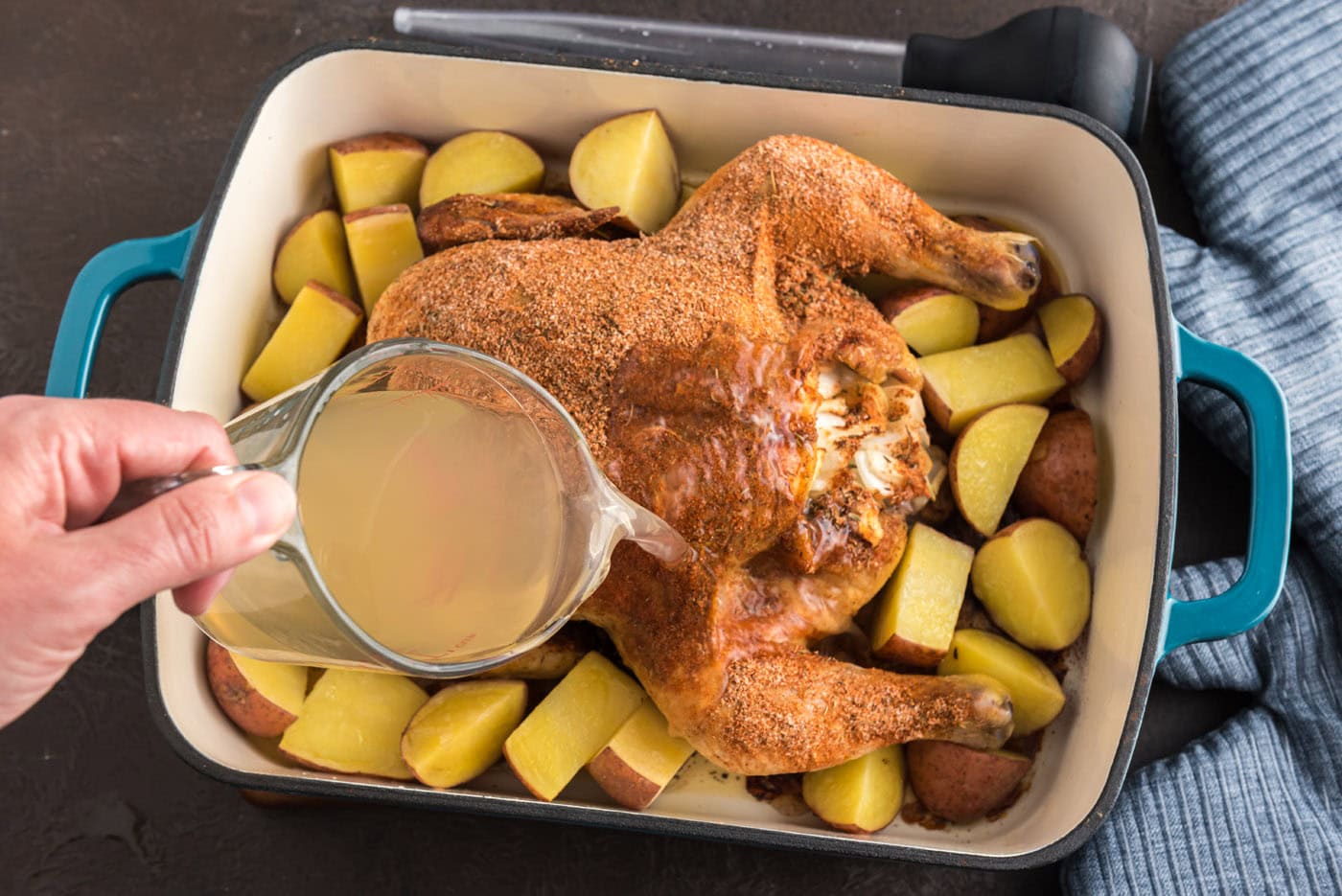 pouring chicken stock over whole roasted chicken in pan with potatoes