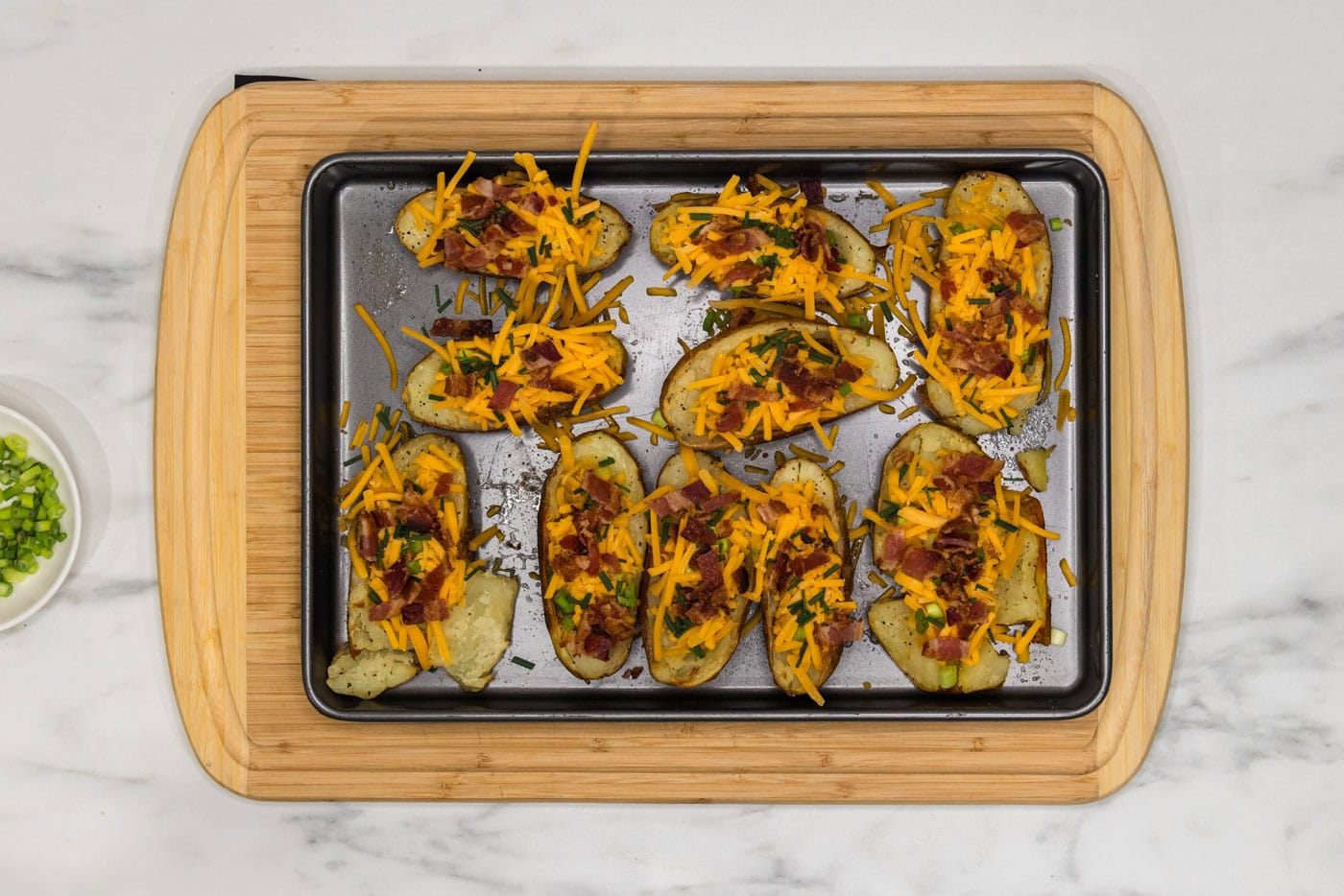 bacon, shredded cheese, chives, and green onions on potato skins on a baking sheet