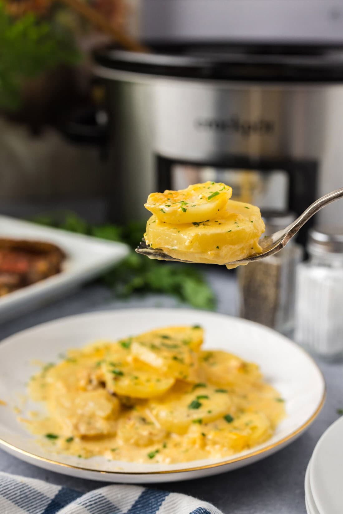 Two Crockpot Scalloped Potatoes on a fork held above a plate of Crockpot Scalloped Potatoes