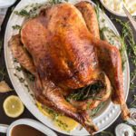 Close up photo of Roasted Turkey on a white serving platter surrounded by herbs and lemon slices