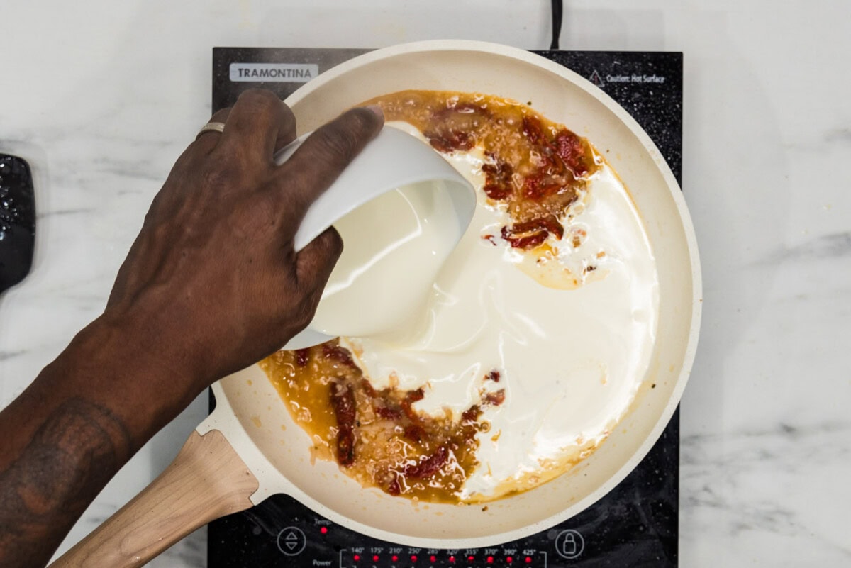 pouring heavy whipping cream into sauce mixture with sun dried tomatoes