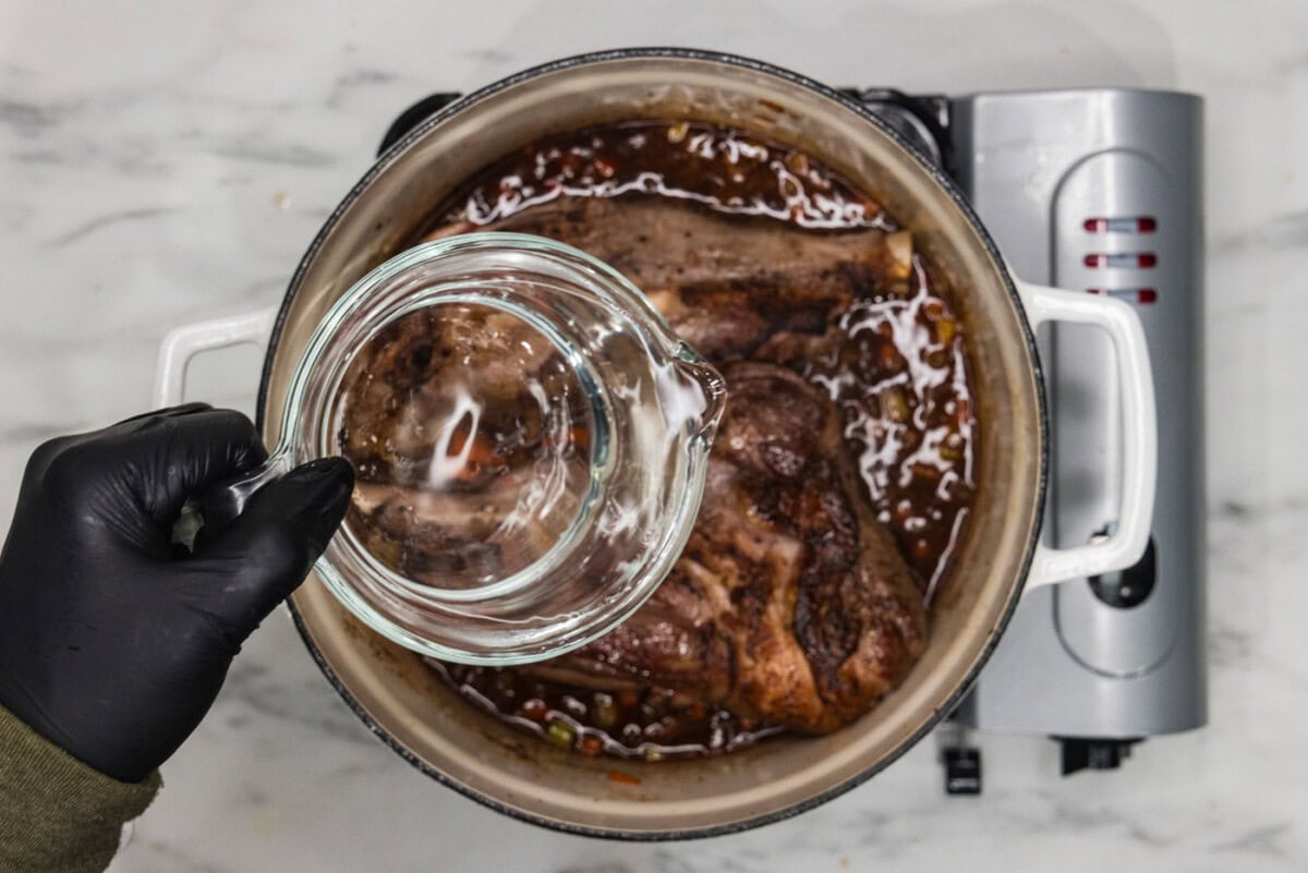 pouring water into dutch oven with braised lamb shanks