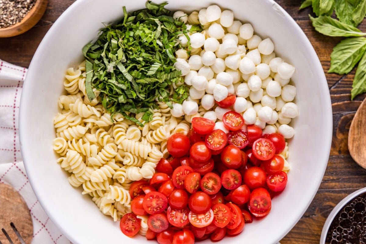 Chopped basil leaves, cherry tomatoes, mozzarella balls, and pasta in a bowl