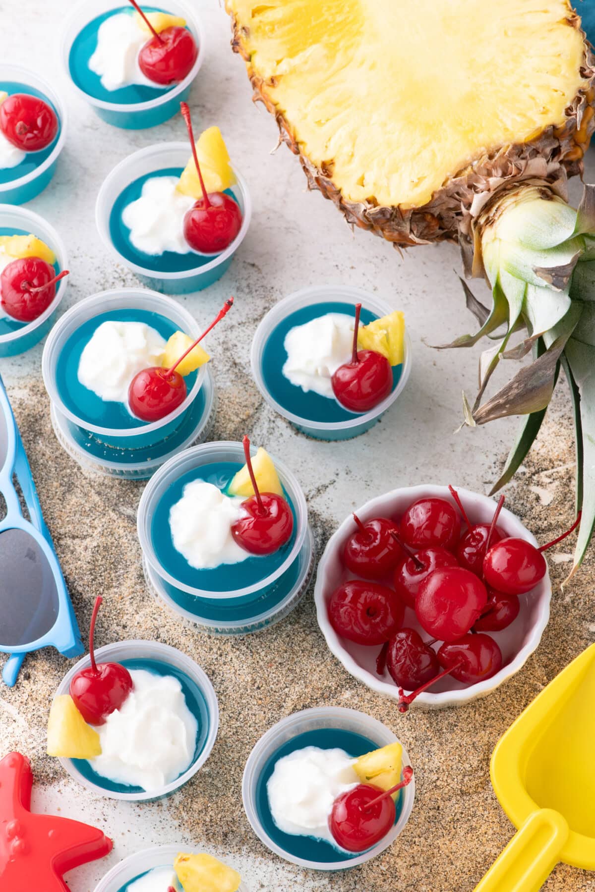 Blue Hawaiian Jello Shots resting on sand with a pineapple and cherries in the background 