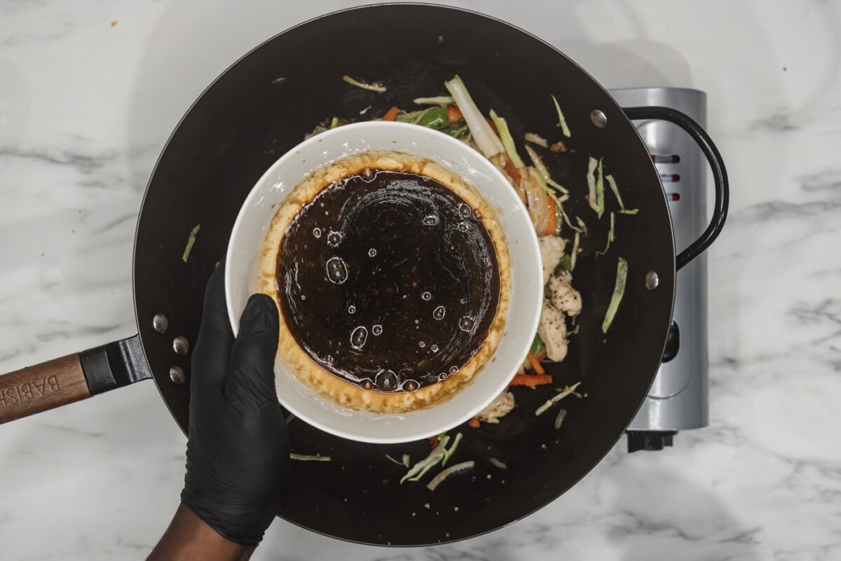 gloved hand holding lo mein sauce in a bowl over a wok