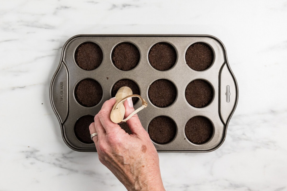 Hand using a pestle to push down graham cracker crust in a mini cheesecake pan
