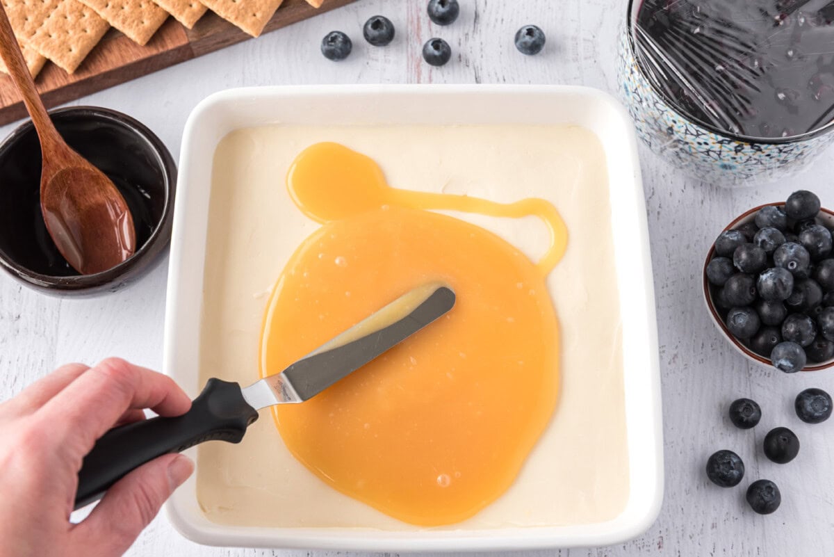 spreading lemon curd over chilled cheesecake in a square pan