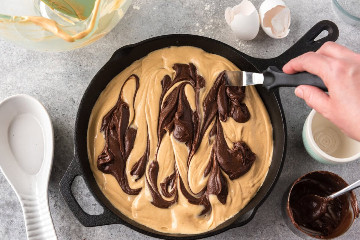 hand using an icing spatula to create swirls of brownie batter on top of peanut butter mixture