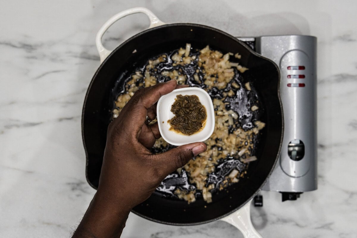 hand holding italian herb base in a small bowl over a skillet of shallots and garlic