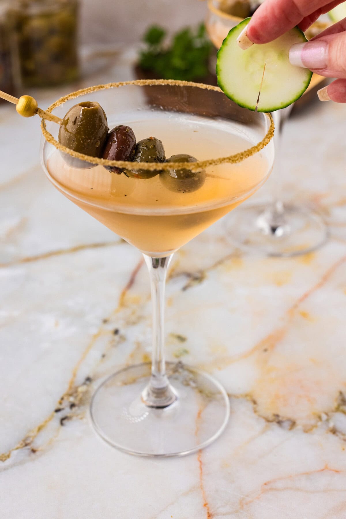 hand adding a cucumber wheel to the side of a dirty greek martini glass