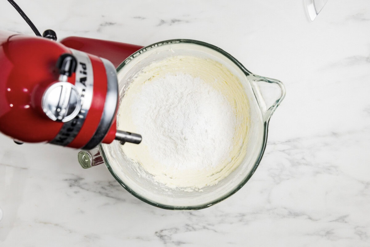 overhead photo of powdered sugar added to beaten butter mixture in a stand mixer
