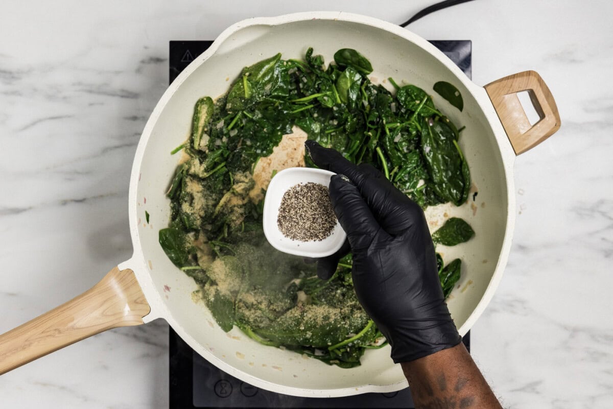 overhead photo of hand adding black pepper to spinach mixture in. skillet