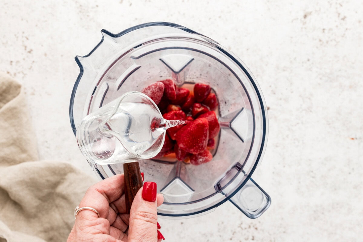 hand pouring silver tequila over top frozen strawberries in a blender