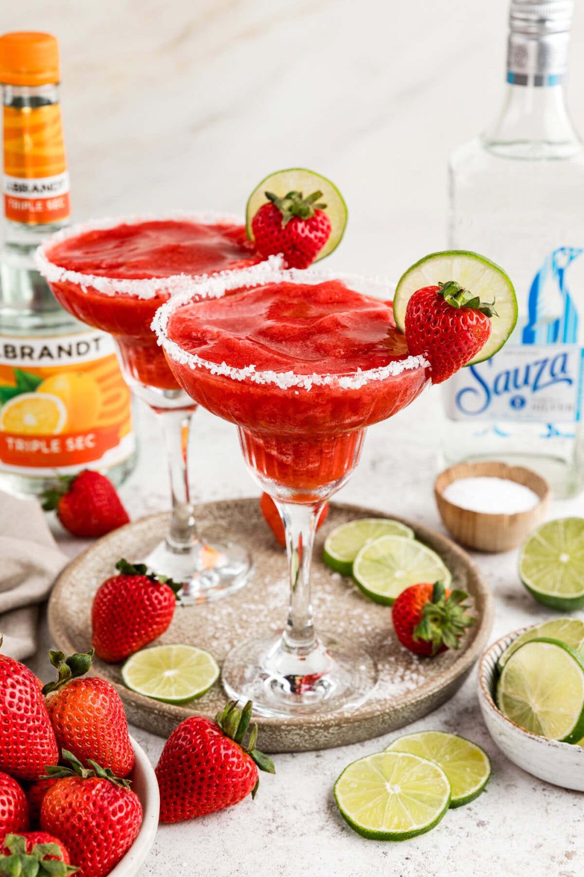 Two Strawberry Margaritas on a serving platter surrounded by limes and strawberries