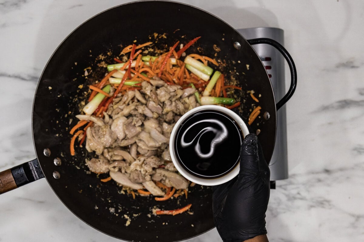 gloved hand holding a bowl of soy sauce over a skillet of chicken and stir fry veggies
