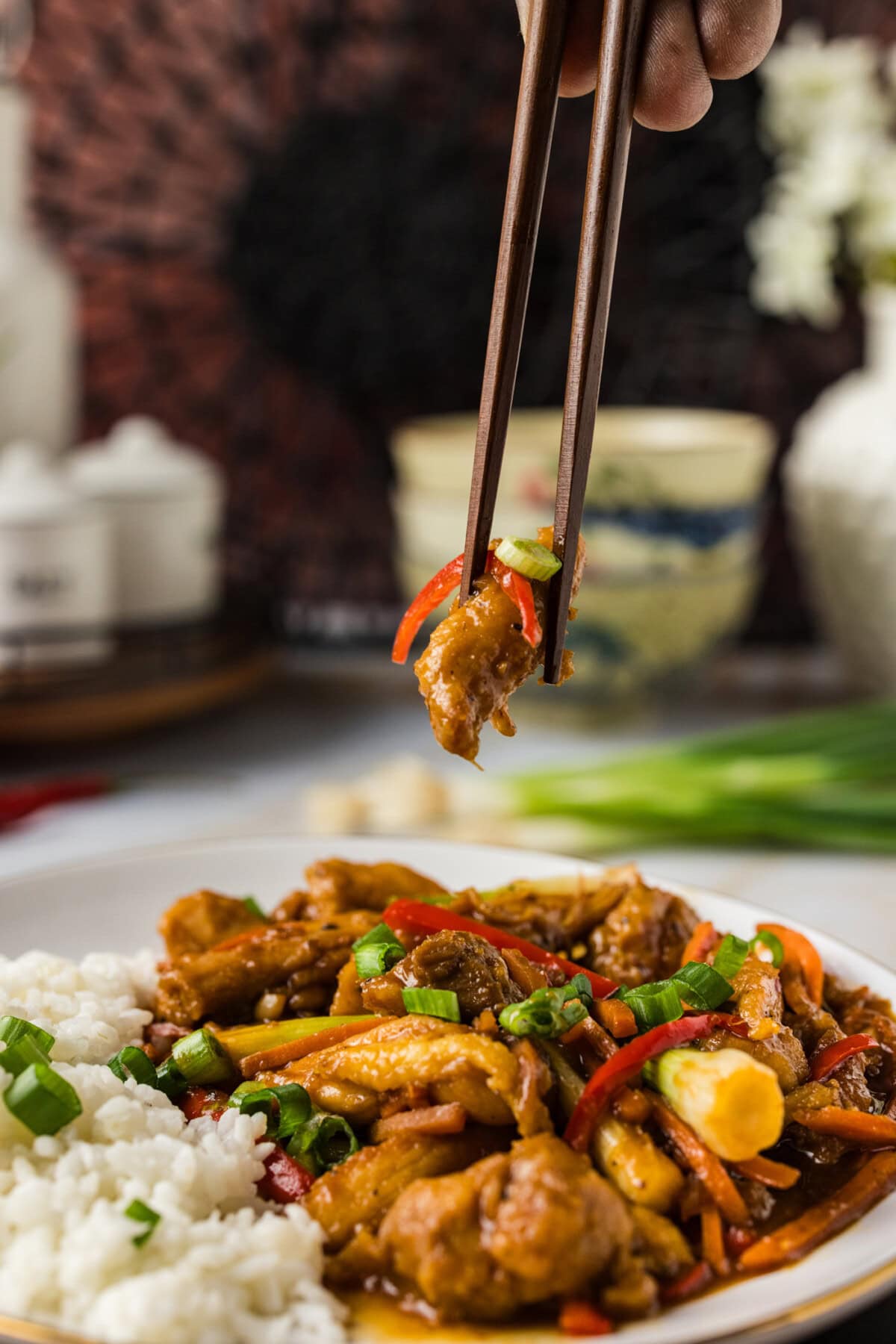 Chopstick holding a piece of Szechuan Chicken over a plate of Szechuan Chicken