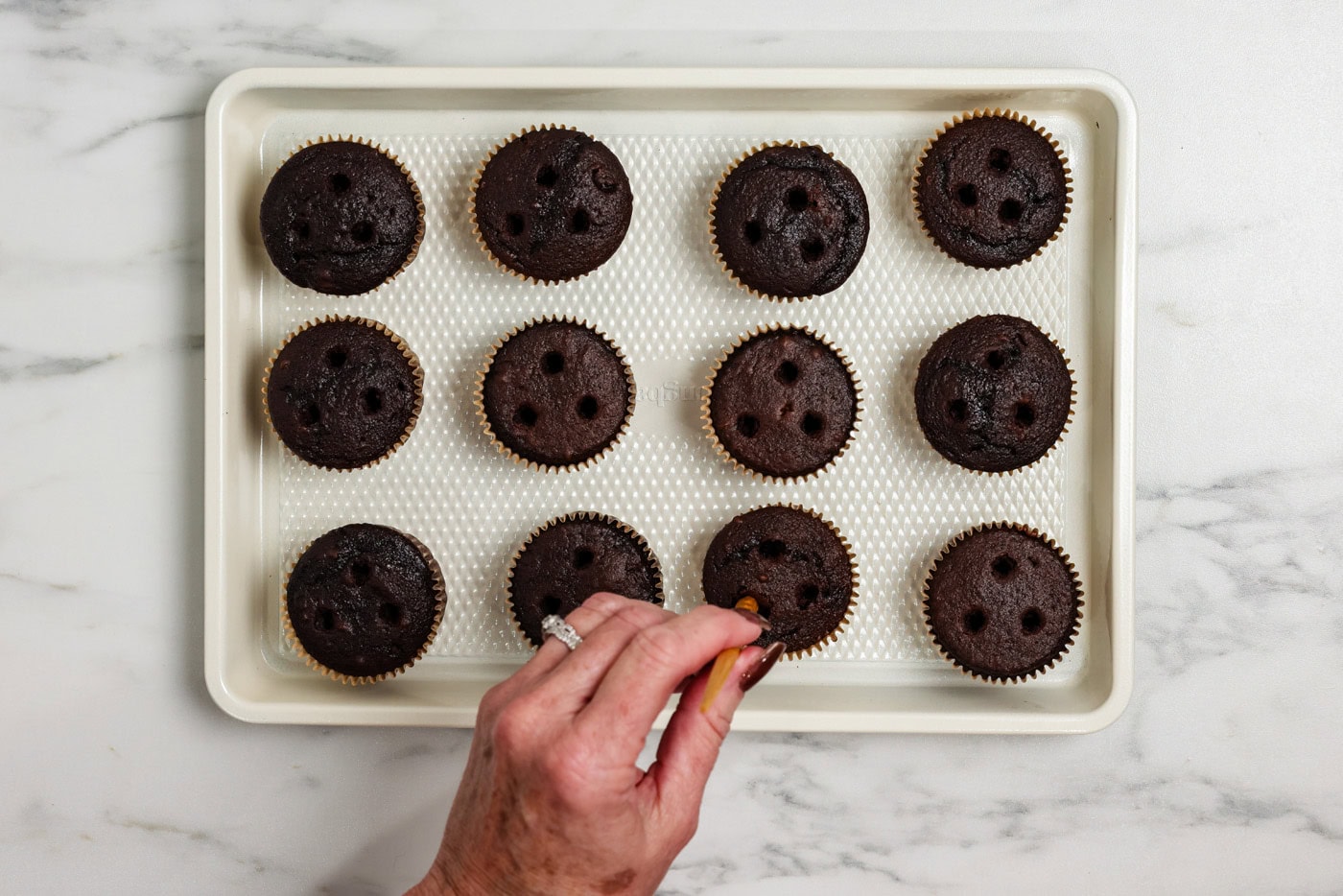 Using a skewer to poke three holes into the top of baked chocolate cupcakes