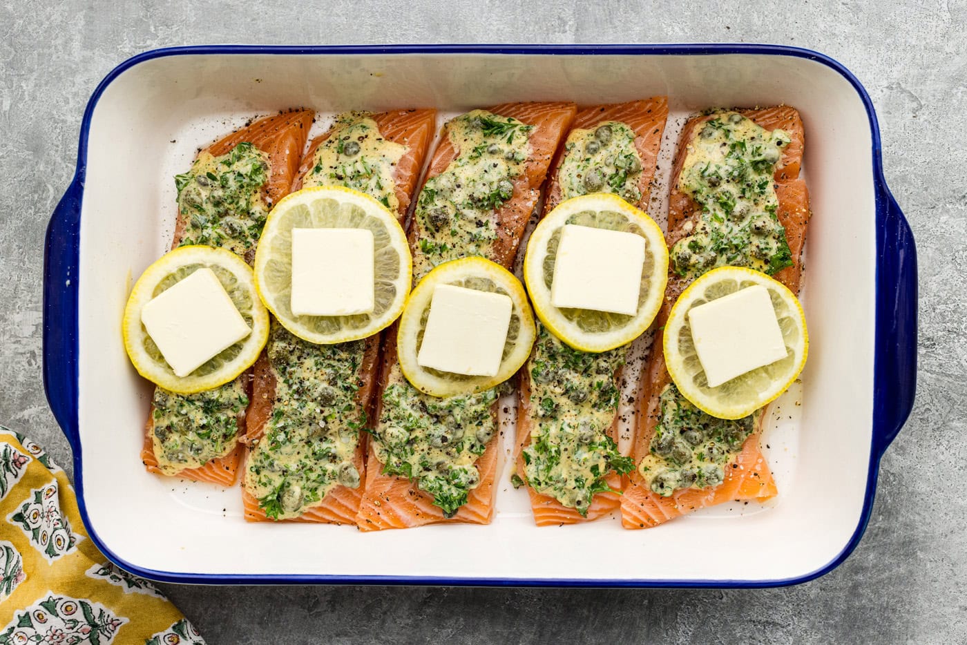 butt pats placed on top of lemon slices over raw lemon caper salmon in a dish