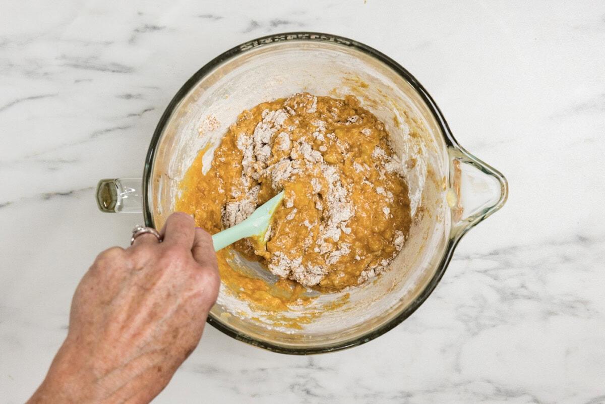 rubber spatula folding in half of the dry ingredients to pumpkin banana batter in a bowl