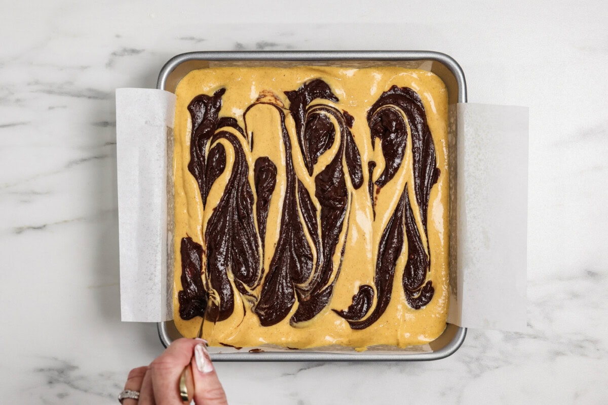 hand using a butter knife to drag spoonfuls of brownie batter over pumpkin cheesecake layer