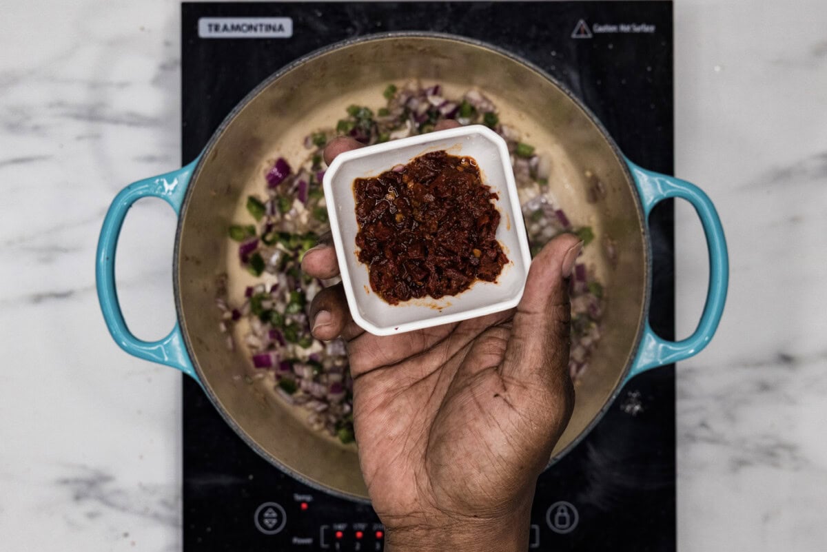 hand holding a bowl of chopped chipotle peppers over dutch oven with sauteed vegetables