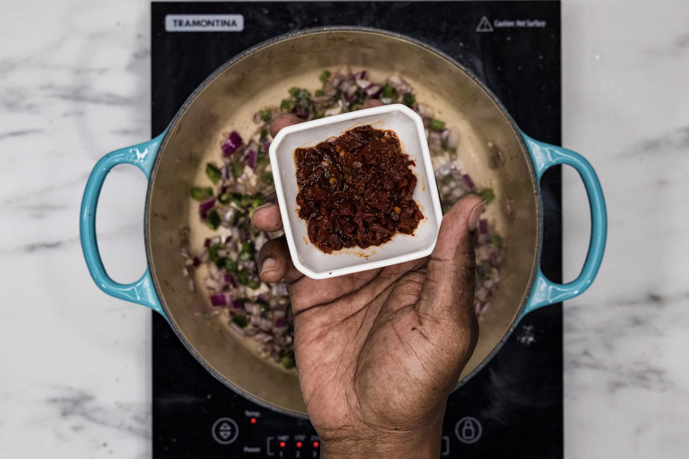 hand holding a bowl of chopped chipotle peppers over dutch oven with sauteed vegetables