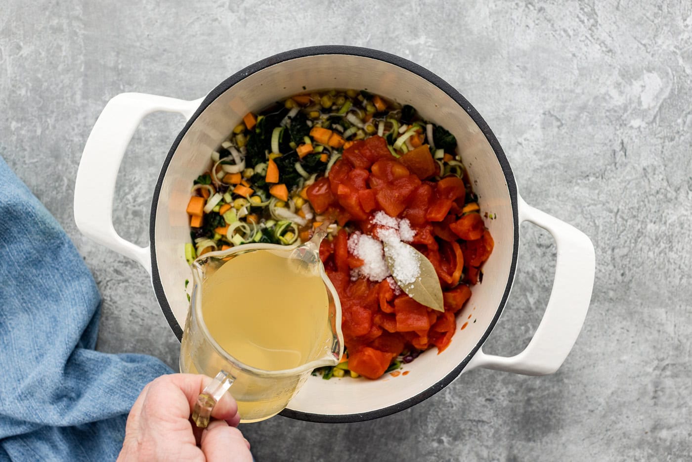 hand pouring vegetable stock into pot with diced tomatoes, bay leaf, and vegetables