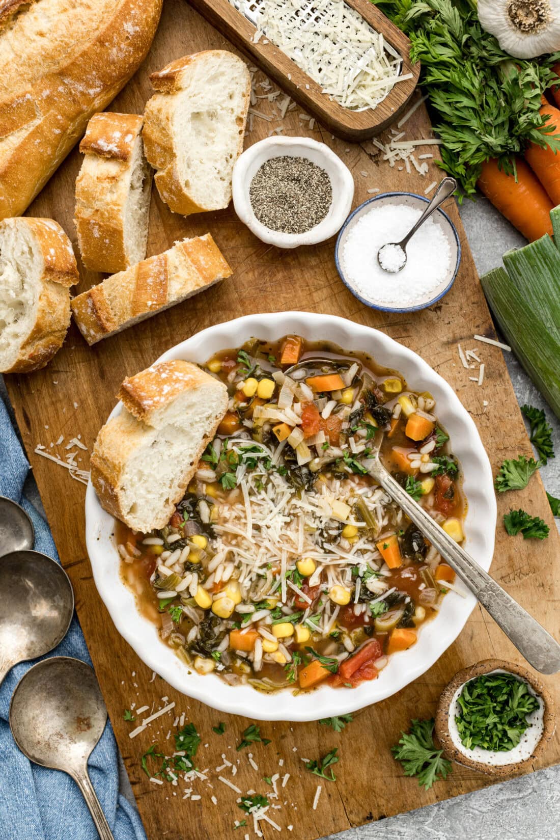 Bowl of Vegetable Rice Soup surrounded by sliced bread, salt, pepper and vegetables