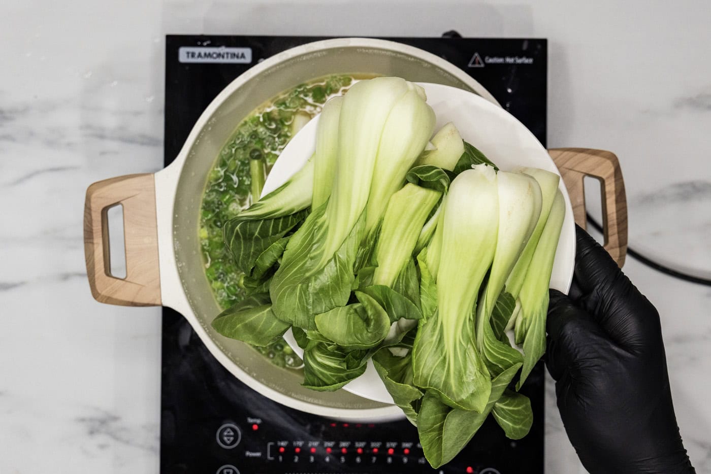 hand adding baby bok choy to stockpot with chicken broth and green onions