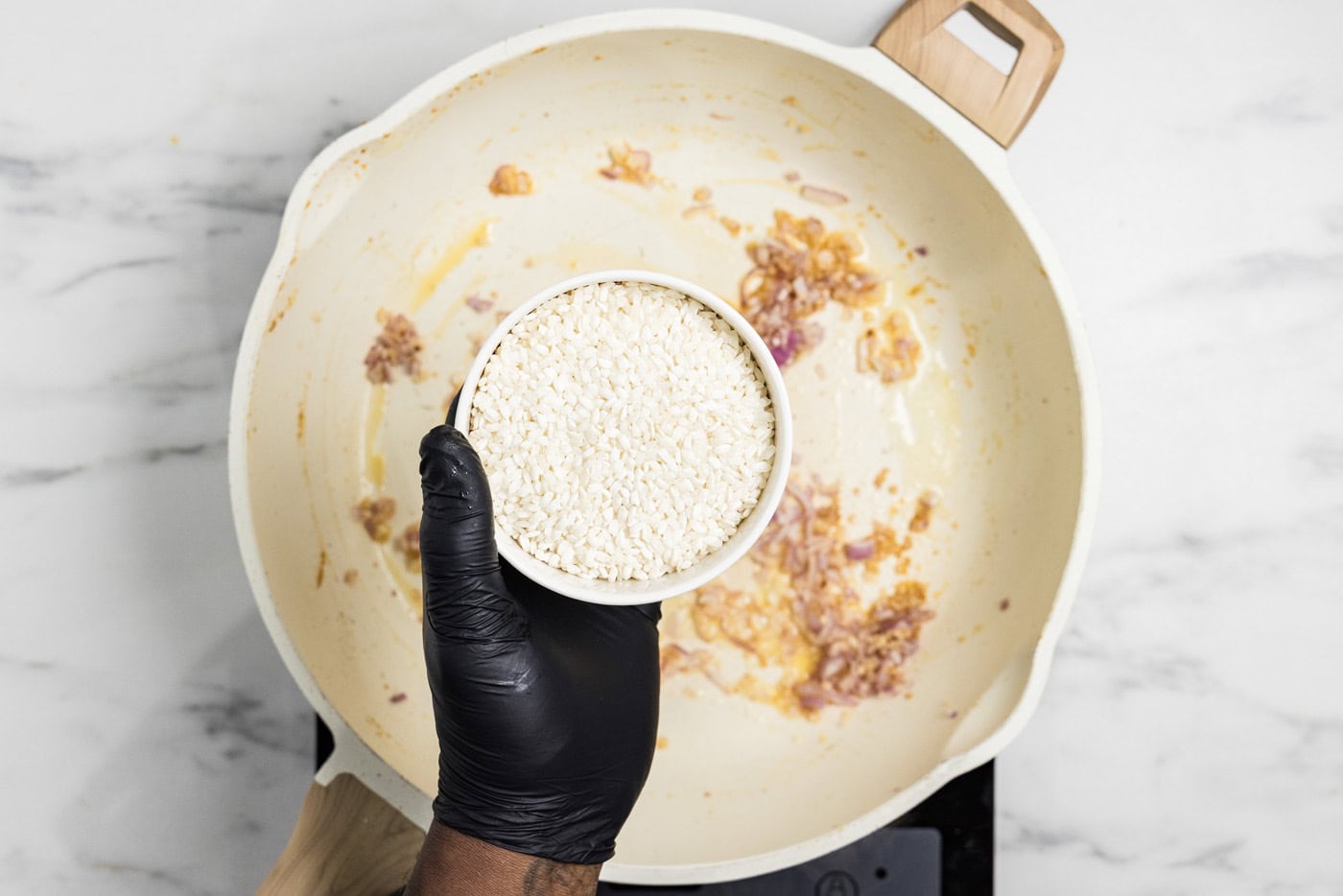 hand holding a bowl of risotto over a skillet with onions and garlic