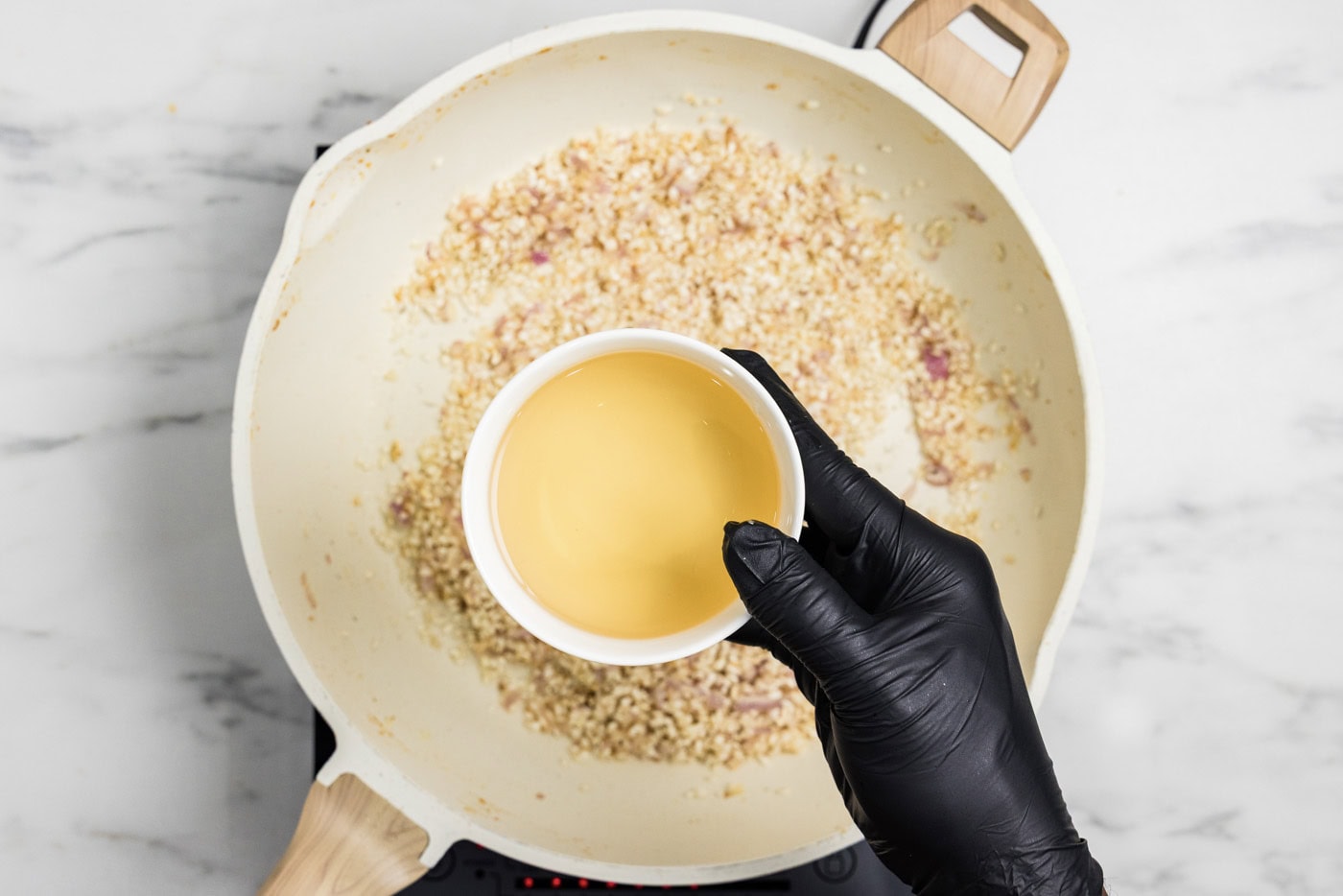 hand holding a bowl of white wine over a skillet of risotto, garlic, and onion