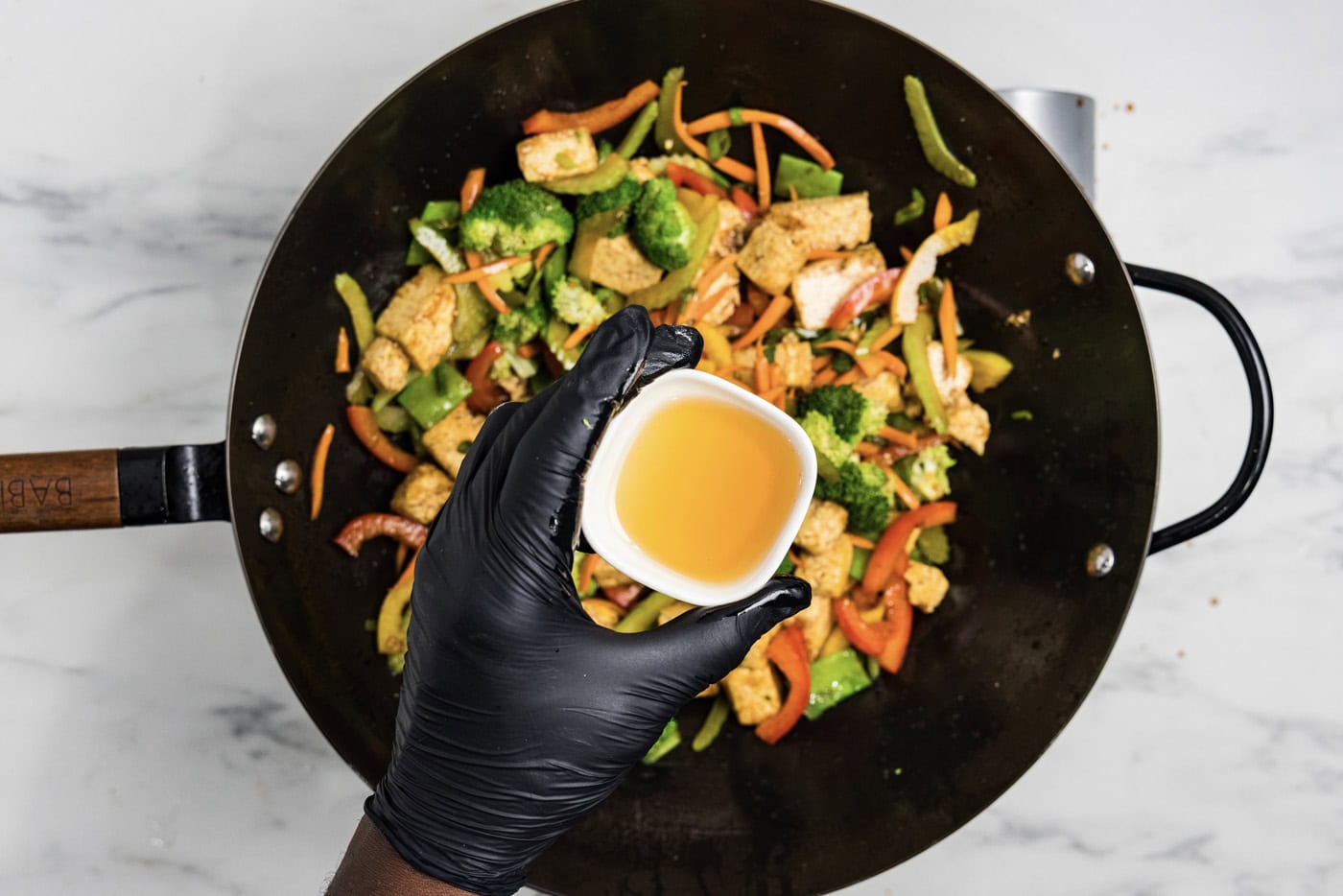 hand holding a small bowl of sesame oil over tofu stir fry in a wok