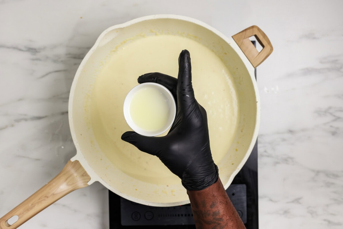 hand holding a small bowl of lemon juice over a skillet of garlic cream sauce