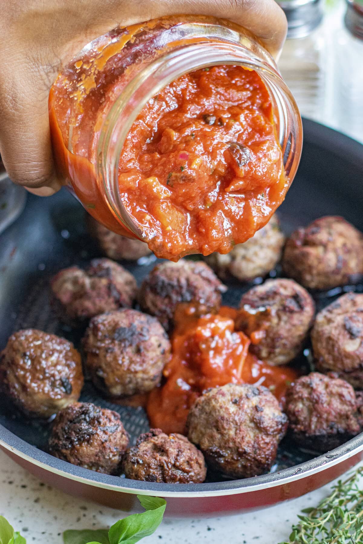 hand pouring jar of marinara sauce over meatballs in a skillet
